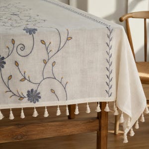 May include: An off-white rectangular tablecloth featuring intricate blue floral embroidery with yellow leaf accents. The fabric has a visible weave texture and is finished with a decorative border of small, hanging off-white fabric tassels.