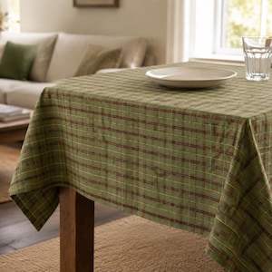 May include: A square tablecloth featuring a green base with a plaid pattern composed of thin intersecting dark red and yellow lines. The fabric drapes over the edges of a wooden table, which holds a single white ceramic plate and a clear glass.