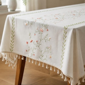 May include: An off-white rectangular tablecloth featuring delicate green vine embroidery and small pink floral accents. The edges are decorated with a repeating green leaf pattern and a border of small, hanging off-white fabric tassels.