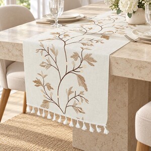 May include: A cream-colored table runner featuring an embroidered brown and tan botanical vine pattern with leaves and small berries. The runner is finished with a row of white fringe tassels along the bottom edge, creating a textured appearance.