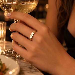 May include: A gold signet ring featuring a flat rectangular face engraved with the name AVA in capital letters. The ring has a polished metallic finish and a smooth band, worn on the finger while holding a clear glass of sparkling liquid.