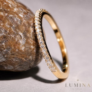 May include: A gold band ring featuring a row of small, clear, round-cut stones set along the outer edge. The ring is positioned next to a smooth, brown, textured stone. The text LUMINA FINE JEWELRY CO is printed in the bottom right corner.