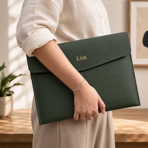 Puede incluir: Una funda para documentos de color verde bosque oscuro con solapa plegable y acabado mate liso. El nombre Lisa está grabado en letras doradas en la solapa frontal. El estuche rectangular presenta bordes cosidos limpios y un perfil plano.