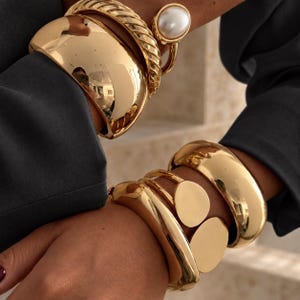 May include: A collection of gold-toned bracelets worn on both wrists. One wrist features a wide, smooth cuff, a twisted rope-style bangle, and a ring with a large white pearl. The other wrist displays a wide cuff and two linked circular discs.