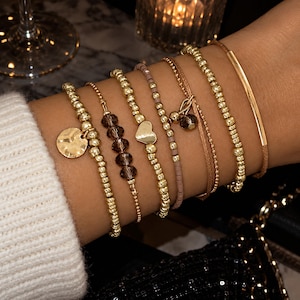 May include: A set of seven stacked bracelets featuring gold-toned metallic beads, a round hammered charm, dark faceted crystal beads, a small gold heart charm, a mauve beaded strand, a thin gold chain, and a smooth gold bar on a brown cord.