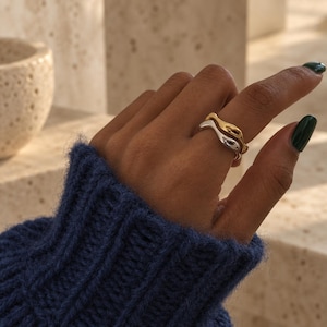 May include: A set of two stacked rings featuring a wavy, organic silhouette. One ring is finished in a polished gold tone, while the other is finished in a polished silver tone. Both bands have a smooth, rounded surface with a fluid, irregular shape.