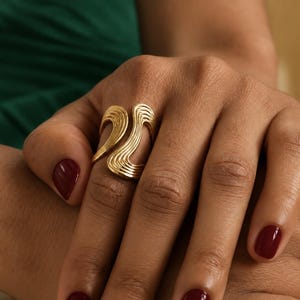 Peut inclure: Une bague dorée dotée d'une large bande ouverte avec une série de lignes ondulées et rainurées parallèles qui créent un aspect fluide et superposé. Le design entoure le doigt avec un espace marqué entre les deux extrémités du métal.
