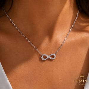 Può includere: Una collana color argento con una catenina sottile e un ciondolo centrale a forma di simbolo dell'infinito, tempestato di piccole pietre chiare e scintillanti. Il gioiello poggia sulla pelle, mostrando un design raffinato e metallico.