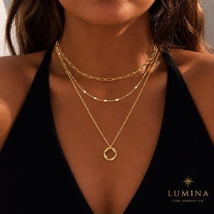 Può includere: Un set di collane dorate a strati composto da tre catene distinte. La catena superiore presenta lunghi maglie ovali, quella centrale è una sottile catena a dischi piatti e la catena inferiore presenta un piccolo ciondolo circolare ritorto.