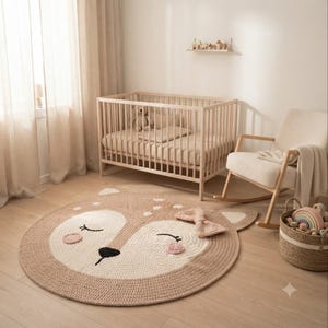May include: A round, crocheted area rug featuring a stylized fawn face with closed eyes, a black nose, and pink cheeks. The rug is tan and cream-colored with a textured bow detail on one side, positioned on a light wood floor in a nursery.