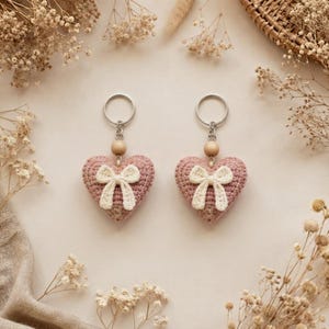 May include: Two heart-shaped keychains made of dusty pink crocheted yarn. Each heart features a small, cream-colored crocheted bow attached to the center and a single round wooden bead positioned above the heart, connected to a silver metal ring.