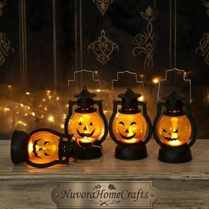 May include: Four black plastic lanterns with orange glass globes featuring black jack-o'-lantern faces. Three lanterns stand upright while one lies on its side. Each lantern has a metal handle and emits a warm yellow light. A metal plaque reads NuvoraHomeCrafts.