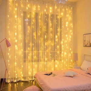 May include: A set of warm white LED curtain lights hanging in vertical strands against a sheer white window curtain. The lights emit a soft glow, creating a bright, illuminated backdrop. The text NuvoraHomeCrafts LED Curtain Lights - 5x6 size is visible.