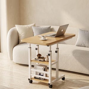 May include: A mobile side table with a light wood-grain top and two lower shelves, supported by a white metal frame with four black caster wheels. The table features adjustable height knobs and holds a laptop, a coffee cup, and books.