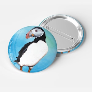 May include: A round button with a colorful illustration of a puffin on a blue background. The puffin is facing to the right and has a black and white body with an orange beak. The button has a silver back with a pin.