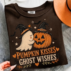 May include: A dark brown t-shirt featuring a graphic of a white ghost and an orange jack-o'-lantern, both wearing black witch hats. The text reads PUMPKIN KISSES GHOST WISHES, surrounded by small stars, hearts, and bats. The label reads COMFORT COLORS.