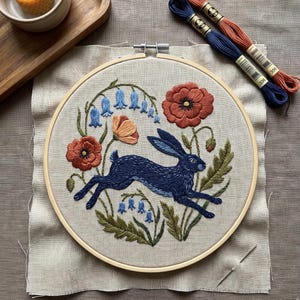 May include: An embroidery hoop containing a fabric panel with a stitched design of a dark blue hare leaping through a field of orange poppies and light blue bell-shaped flowers. Two skeins of embroidery floss in blue and orange sit nearby.