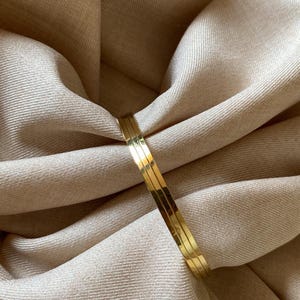 Può includere: Un bracciale rigido color oro lucido caratterizzato da una superficie liscia e riflettente con un sottile dettaglio a scanalatura centrale che corre lungo l'intera circonferenza della fascia circolare, creando un aspetto pulito e uniforme.