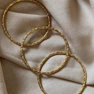Può includere: Tre bracciali rigidi color oro caratterizzati da una texture intrecciata simile a una corda. Ogni bracciale circolare presenta una finitura lucida e riflettente, con uno spessore costante che conferisce un aspetto uniforme a tutti i pezzi.
