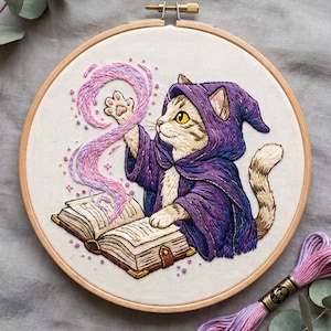 May include: A circular wooden embroidery hoop containing a fabric panel with a hand-stitched design of a tabby cat wearing a purple hooded robe. The cat is casting a pink and purple swirl of magic from an open book with gold-trimmed pages.