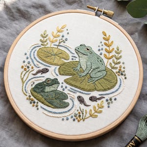 May include: A circular wooden embroidery hoop containing white fabric with a hand-stitched design of two green frogs sitting on lily pads, surrounded by small tadpoles, blue water ripples, and yellow leafy sprigs, with embroidery thread nearby.