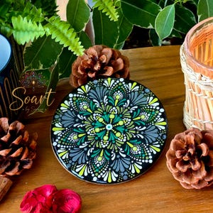 Peut inclure: Un dessous de verre circulaire présentant un motif de mandala peint par points détaillé dans des tons de vert citron, vert forêt, blanc et gris anthracite sur un fond noir. L'œuvre affiche des motifs symétriques de points.
