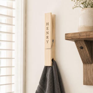 May include: A large, light-coloured wooden clothespin wall hook featuring the name HENRY engraved vertically in black capital letters. The functional clip mechanism includes a silver-toned metal spring, and a dark grey towel hangs from the bottom.