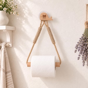 May include: A wall-mounted toilet paper holder featuring a light wood whale-shaped wall mount with the name Oliver engraved on it. Two thick jute ropes hang from the whale, supporting a light wood dowel that holds a roll of white toilet paper.