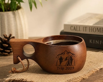 May include: A dark brown wooden cup with a handle featuring a circular cutout and a leather cord. The exterior is engraved with a mountain and forest graphic above the text Trip 2025. The cup is filled with a dark liquid and has a smooth finish.