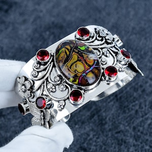Op de afbeelding: Een zilveren manchetarmband met een grote, ovale, iriserende steen in het midden. De metalen band is versierd met gedetailleerde bloem- en vlindermotieven en is geaccentueerd met verschillende kleine, ronde, gefacetteerde rode granaatstenen.