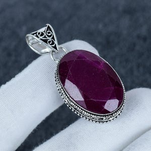 Op de afbeelding: Een ovaalvormige, gefacetteerde dieprode edelsteen hanger gezet in een decoratieve zilveren omlijsting. De hanger heeft een driehoekig oogje met ingewikkelde krulpatronen en een kralenrand rondom de centrale zetting van de steen.