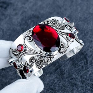 Puede incluir: Un brazalete de plata tipo brazalete con una gran piedra preciosa roja oscura facetada de forma ovalada en el centro. La superficie metálica está decorada con intrincados relieves de volutas y hojas, acentuados por seis piedras rojas redondas.