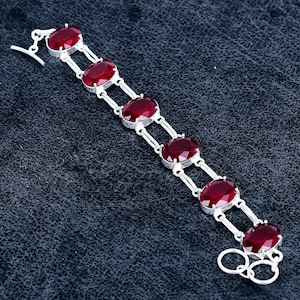 May include: A silver-toned bracelet featuring six oval-shaped, deep red faceted stones set in a double-link chain design. The bracelet includes a toggle clasp closure with a T-bar and a circular ring at the end, showcasing a polished metallic finish.