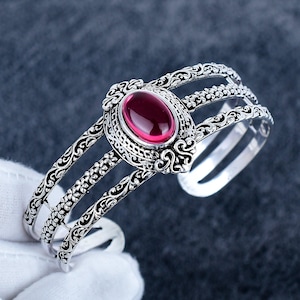 Può includere: Un bracciale a fascia color argento con tre bande parallele caratterizzate da intricati motivi floreali e a volute. Il centro del bracciale è decorato con una grande pietra cabochon ovale rosso scuro incastonata in una cornice in filigrana.