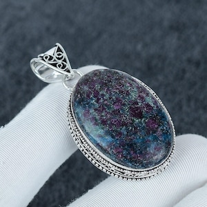 Peut inclure: Un pendentif de forme ovale présentant une pierre polie multicolore avec des inclusions magenta profond et bleu-vert foncé. La pierre est sertie dans une monture en argent sterling décorative avec une bélière en filigrane détaillée.