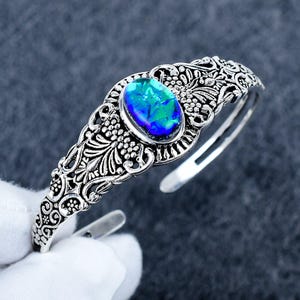 May include: A silver-toned cuff bracelet featuring an oval-shaped iridescent blue and green stone at the center. The metal band displays intricate filigree scrollwork and floral patterns throughout, with an open back design for an adjustable fit.