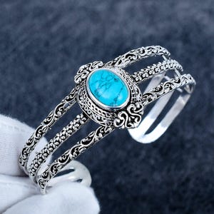 May include: A silver-toned cuff bracelet featuring an oval-shaped turquoise stone with dark veining at the centre. The band consists of three parallel metal strands decorated with intricate scrollwork and floral patterns throughout the design.
