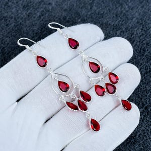 May include: A pair of long chandelier earrings featuring multiple deep red, pear-shaped gemstones set in polished silver-toned metal. Each earring consists of a central teardrop frame adorned with several dangling red stones suspended from a hook.