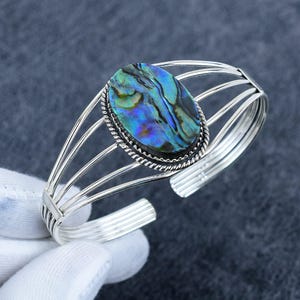 Può includere: Un bracciale a fascia con un inserto centrale ovale in conchiglia di abalone dai toni blu e verdi iridescenti. La conchiglia è incorniciata da un bordo in metallo intrecciato e montata su una fascia a più fili composta da cinque fili argentati.