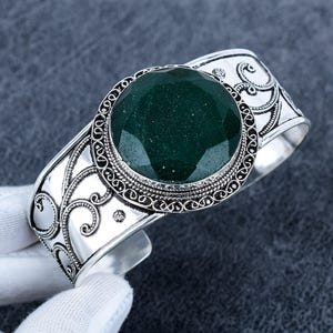 Puede incluir: Un brazalete de plata con una gran piedra preciosa redonda y facetada de color verde oscuro en el centro. La banda de metal está decorada con intrincados patrones de volutas en relieve y un borde de cuentas que rodea el engaste central.