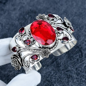 Puede incluir: Un brazalete tipo puño de tono plateado con una gran piedra preciosa roja de corte ovalado en el centro. La superficie metálica está decorada con intrincados arabescos, motivos florales y varias piedras rojas pequeñas en forma de lágrima.