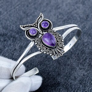 Peut inclure: Un bracelet manchette en argent avec un motif central en forme de hibou. Le design du hibou est orné de volutes métalliques complexes et serti de deux pierres d'améthyste violettes rondes pour les yeux et d'une en forme de goutte pour le corps.
