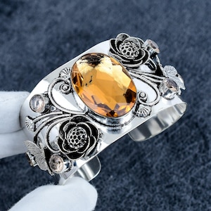Może przedstawiać: Srebrna bransoletka typu mankiet z dużym, owalnym, fasetowanym kamieniem w kolorze bursztynowym pośrodku. Metalowe zdobienia obejmują misterne motywy kwiatowe z ciemnymi, oksydowanymi detalami oraz kilka mniejszych, okrągłych, przezroczystych kamieni.