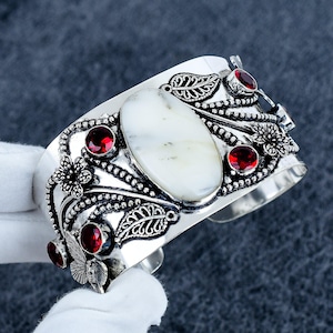 Può includere: Un bracciale a fascia in argento con una grande pietra ovale bianca centrale, circondata da intricate lavorazioni in metallo a forma di foglie, fiori e farfalle. Il design è arricchito da diverse pietre rotonde sfaccettate rosso scuro.