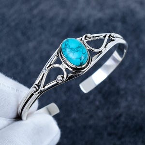 May include: A silver cuff bracelet featuring an oval-shaped turquoise stone with dark veining at the centre. The metal band displays an openwork design with swirling patterns and small spherical accents, creating a decorative frame around the stone.