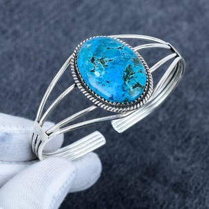 May include: A silver cuff bracelet featuring a large, oval-shaped turquoise stone with dark matrix veining. The stone is set in a twisted rope border, and the band consists of four polished silver wires that converge at the sides of the setting.