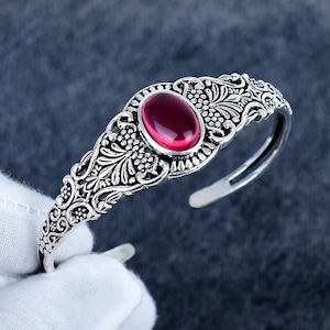 May include: An open cuff bracelet crafted from silver-toned metal featuring an intricate filigree pattern with floral and vine motifs. A smooth, oval-shaped deep red cabochon gemstone is set prominently in the center of the decorative metalwork.