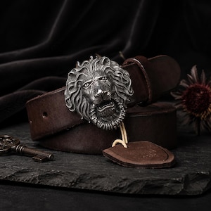 Può includere: Una cintura in pelle marrone scuro con una fibbia in metallo color argento a forma di testa di leone dettagliata. La cintura include un'etichetta in pelle abbinata fissata con un cordino e la fibbia presenta trame complesse nella criniera.