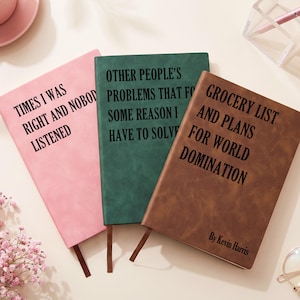 Può includere: Tre quaderni rettangolari rosa, verde scuro e marrone con testo nero. Il quaderno rosa riporta: TIMES I WAS RIGHT AND NOBODY LISTENED. Quello verde: OTHER PEOPLE'S PROBLEMS THAT FOR SOME REASON I HAVE TO SOLVE. Quello marrone: GROCERY LIST AND PLANS FOR WORLD DOMINATION By Kevin Harris.