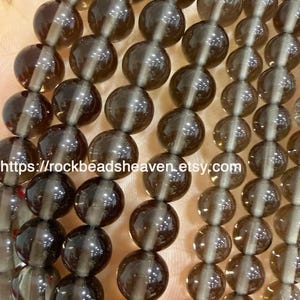May include: Multiple strands of round, translucent, smoky brown quartz beads strung on thin cord. The beads have a smooth, polished finish and a consistent spherical shape. The text https://rockbeadsheaven.etsy.com is visible.